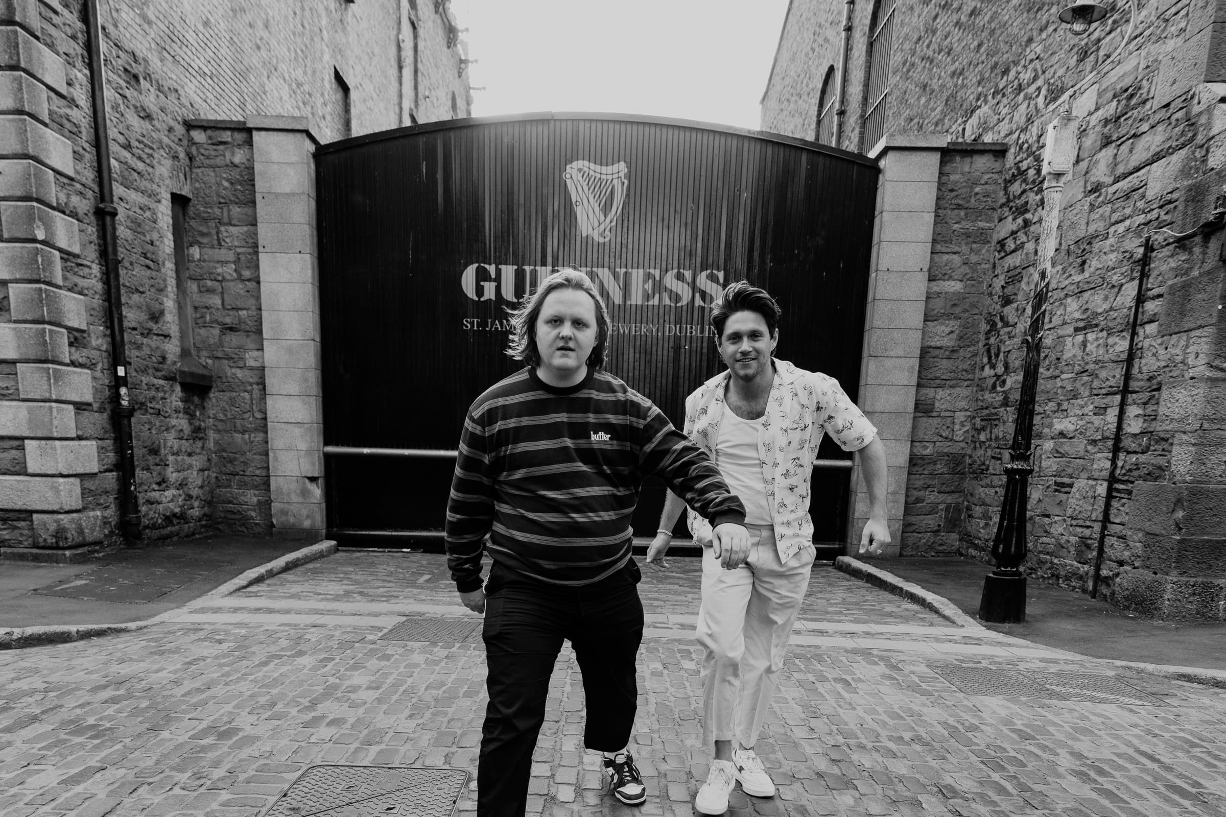 Two men stride over cobbles towards the camera in front of the Guinness brewery gate